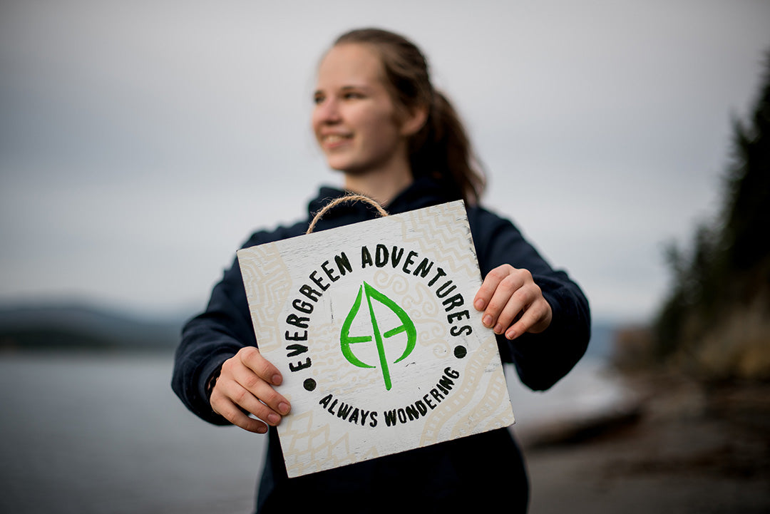 Evergreen Adventures owner Rainee is holding a wood board with the Evergreen Adventures leaf logo painted on it