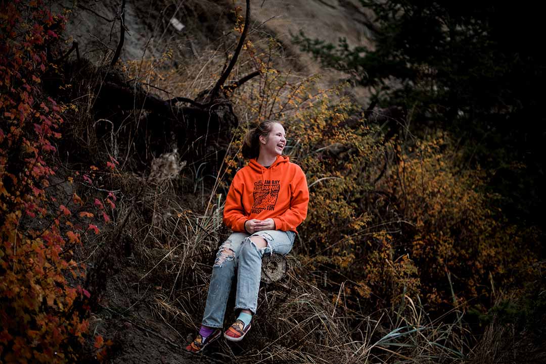 Person (Rainee) Wearing a bright orange hoodie with a Clallam Bay design on it sitting in a log laughing and surronded by fall leaves